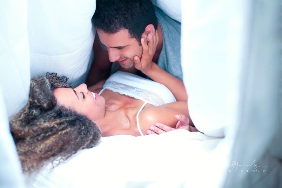 couple smiling at each other laying on bed under sheets