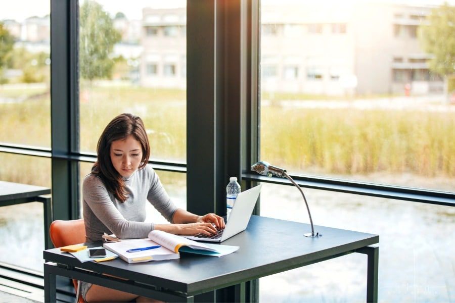 college student taking notes from books in library as she studies