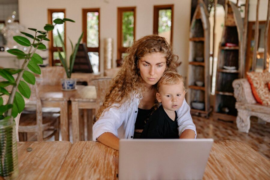 Mother and child sit at a wooden table with a laptop indoors, focusing together.