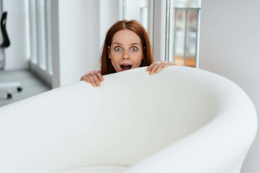 Surprised young woman peeking over a large white tub chair with open mouth and wide eyes indoors