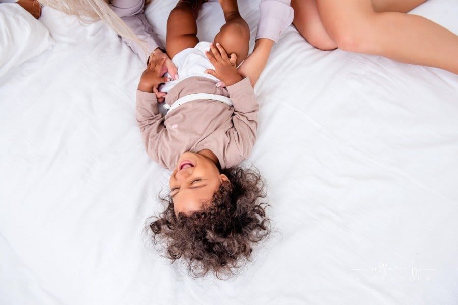 mother tickles her little daughter while lying on a white bed, having fun, top view.