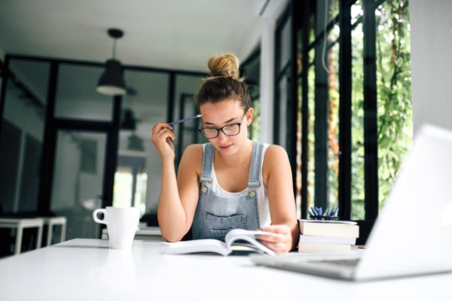 young woman wearing overalls reading a book while sitting in front of online learning class
