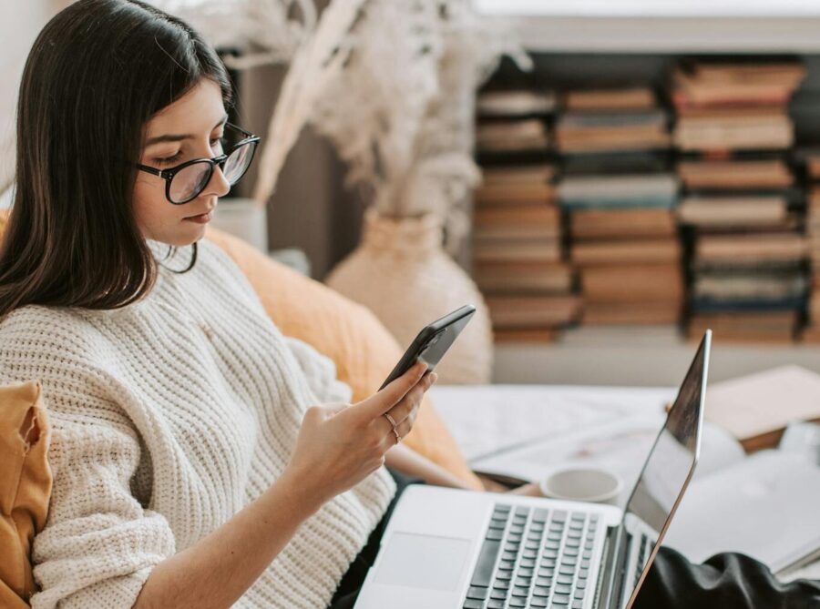 Side view of focused young female student sitting on bed at home while browsing Internet on laptop and using smartphone