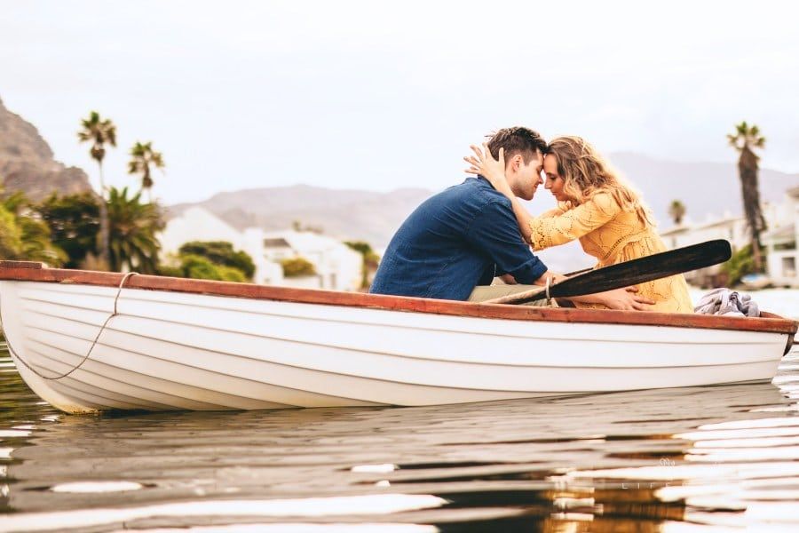Young couple sitting together in a boat touching their heads. Couple in love on a boat date in a lake with hills and houses i