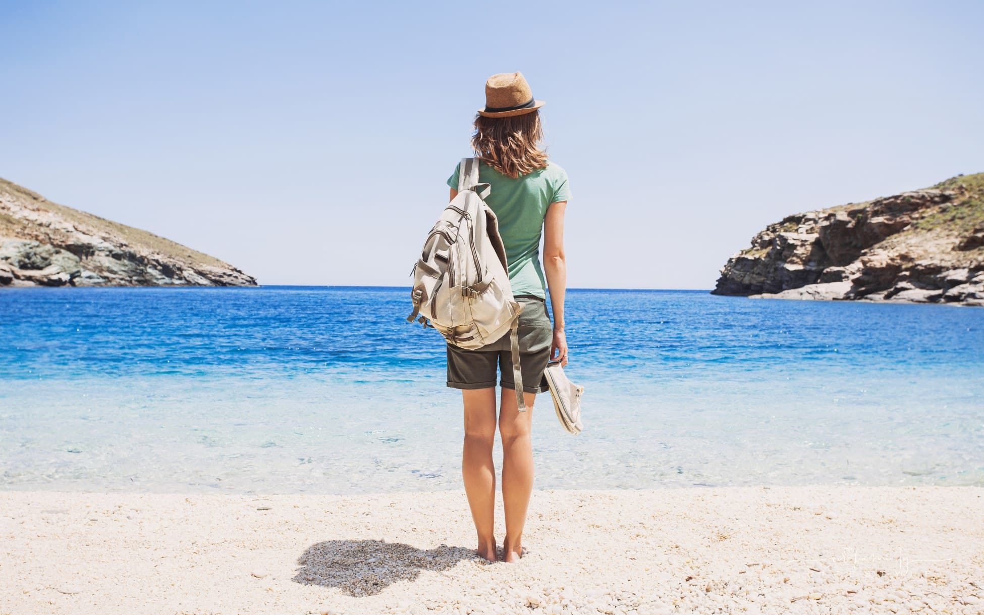 female traveler loking over ocean while standing on beach with shoes in hand and backpack on