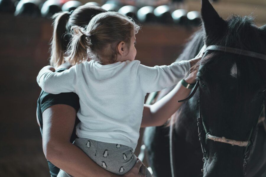 Woman holding child petting a horse at a stable, showcasing gentle bonding and animal interaction.