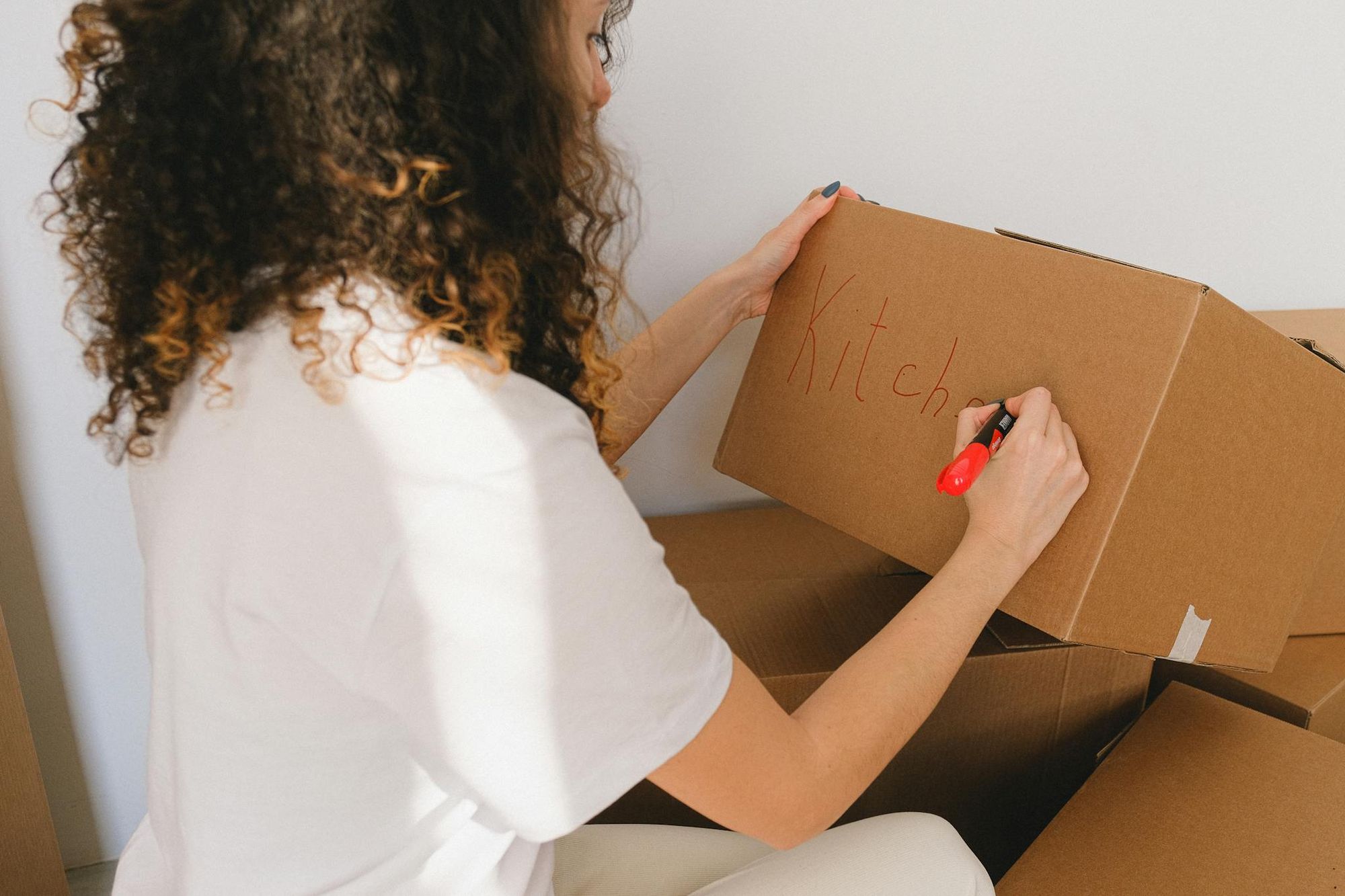 Woman writing 'Kitchen' on a cardboard box with a red marker, preparing for moving.