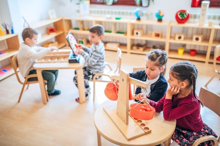 Two preschool age girls sitting together in their classroom and playing with scale. There are other school boys playing in th