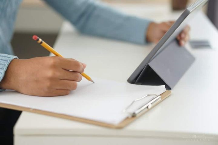 male student writing on paper notebook while looking at digital tablet