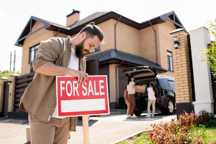 bearded man in casual shirt placing For sale sign against house to pay attention of buyers