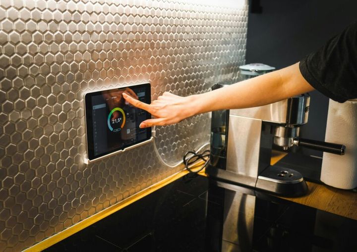 Modern kitchen scene featuring a hand using a tablet to control a smart home system.