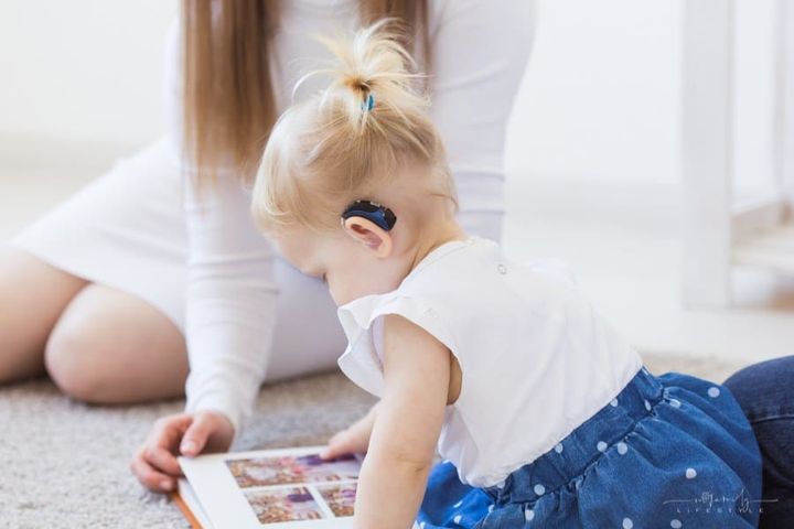 toddler girl wearing a hearing aid while looking at book with mom