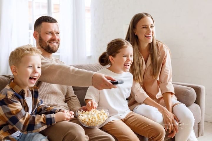 Delighted family parents and kids eating popcorn and laughing while sitting on sofa and watching funny movie at home