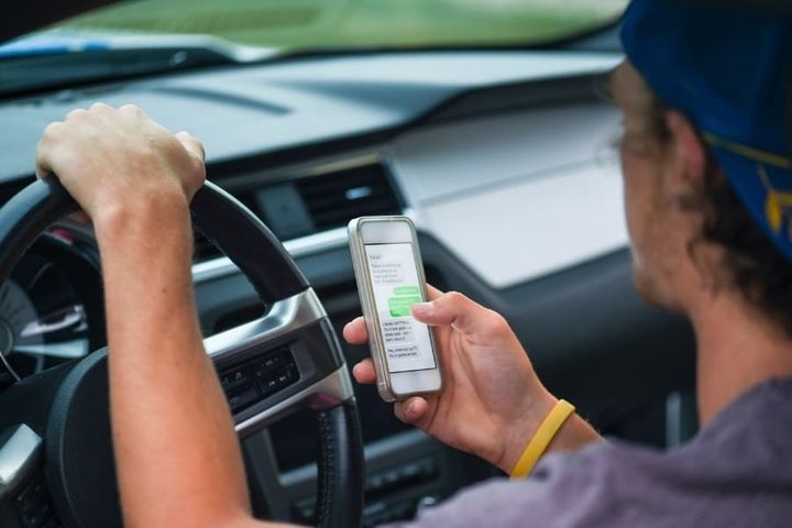 A young man drives with one hand on the steering wheel while dangerously texting on a mobile phone in the other hand.