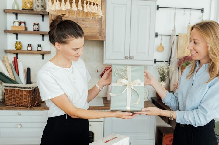 Two women in a home office exchanging a beautifully wrapped gift, symbolizing friendship and celebration.