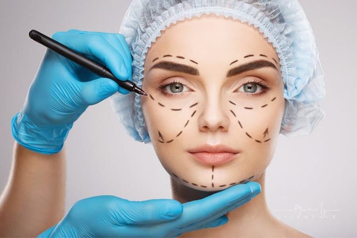 Pretty girl with dark eyebrows wearing blue medical hat at studio background, doctor's hands wearing blue gloves drawing perf