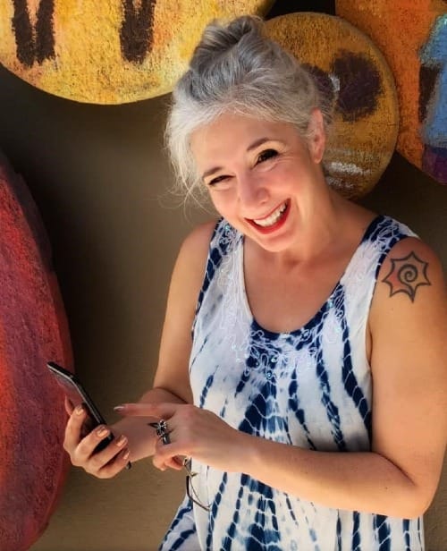 Roxanne Lynch smiling in a blue-and-white dress while holding a phone, representing confidence, visibility, and using Facebook keyword strategy to build online income from home.