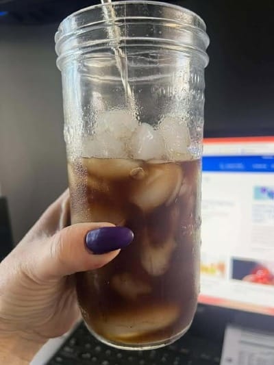 Hand holding a cold mushroom blend drink in front of a computer screen, symbolizing calm focus and natural energy support while working from home.