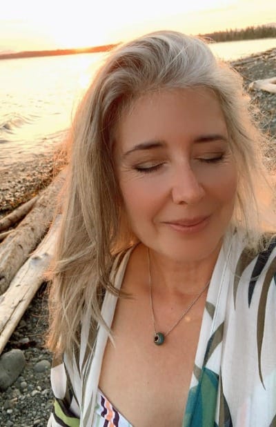 Woman standing by the water at sunset with eyes closed, symbolizing calm confidence, emotional strength and steady energy while building a life and income from home.