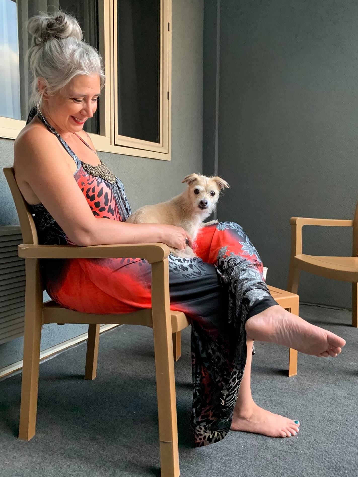 Roxane Lynch relaxing barefoot on a patio chair with her small dog on her lap, smiling peacefully during a quiet moment of reflection and freedom.