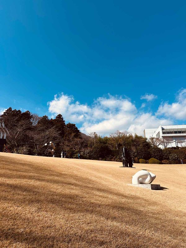 Hakone Open Air Museum: a piercingly blue sky with some clouds over a yellowed grassy hill with a few sculptures, some dark trees, and a white building
