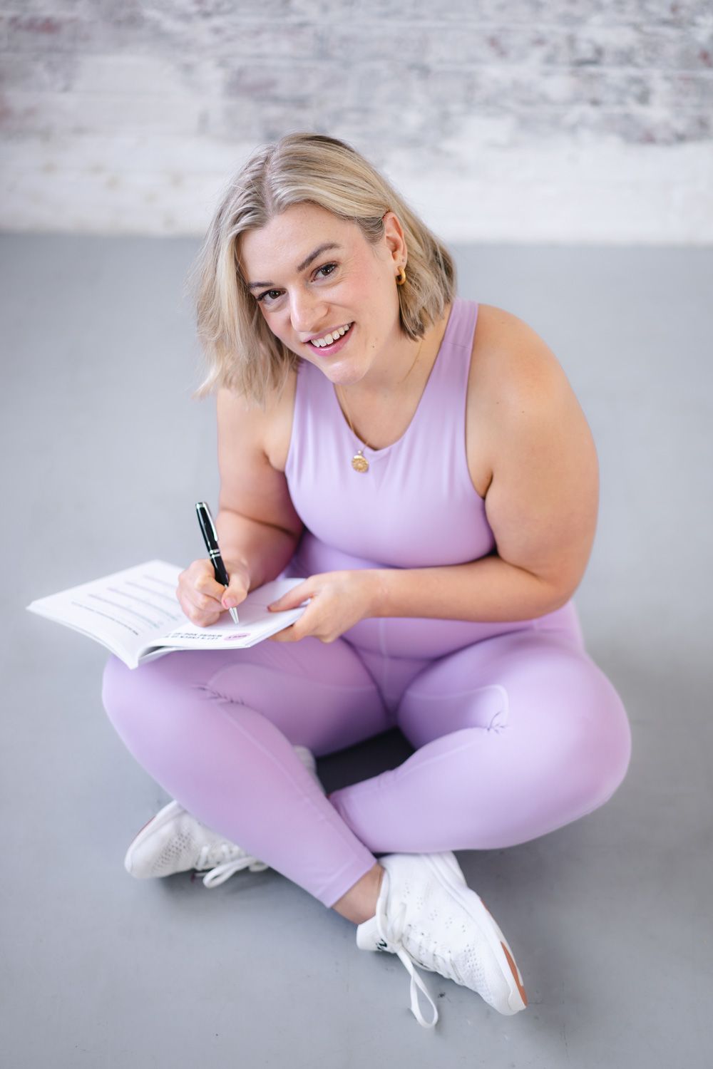 Tally Rye is sitting wearing exercise clothes and filling in her journal, she is smiling at the camera