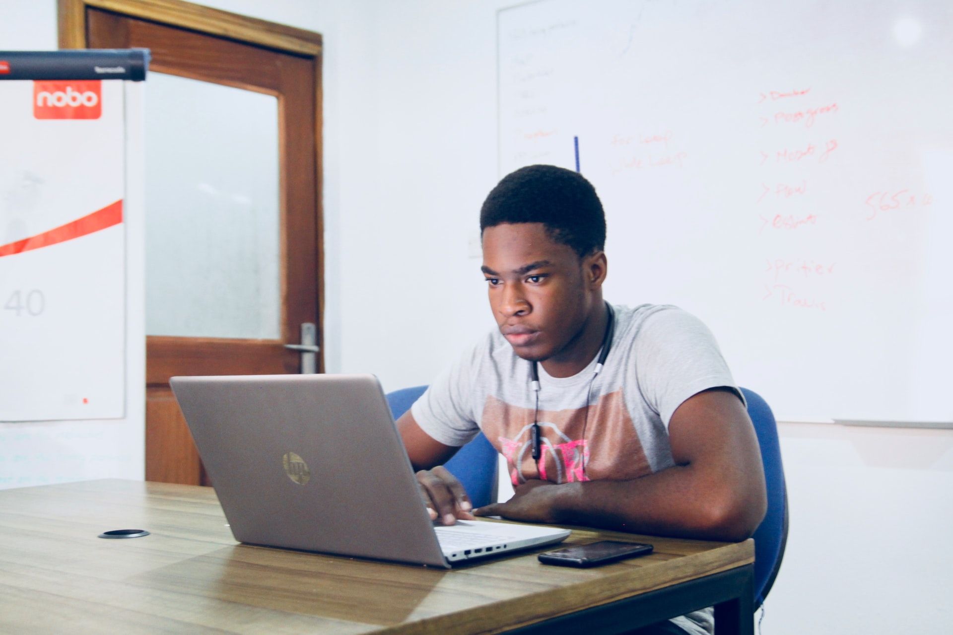 A young man sits alone in a classroom, looking at his laptop.