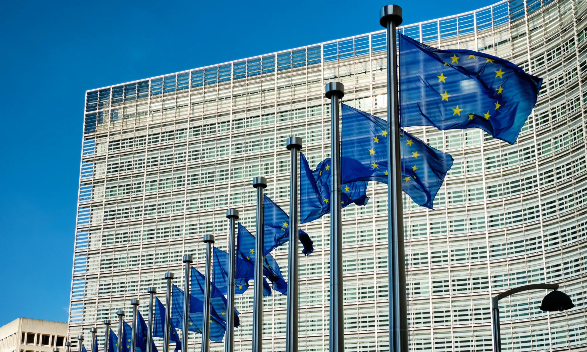 European Union flags waving in front of the modern glass-fronted European Commission building in Brussels.