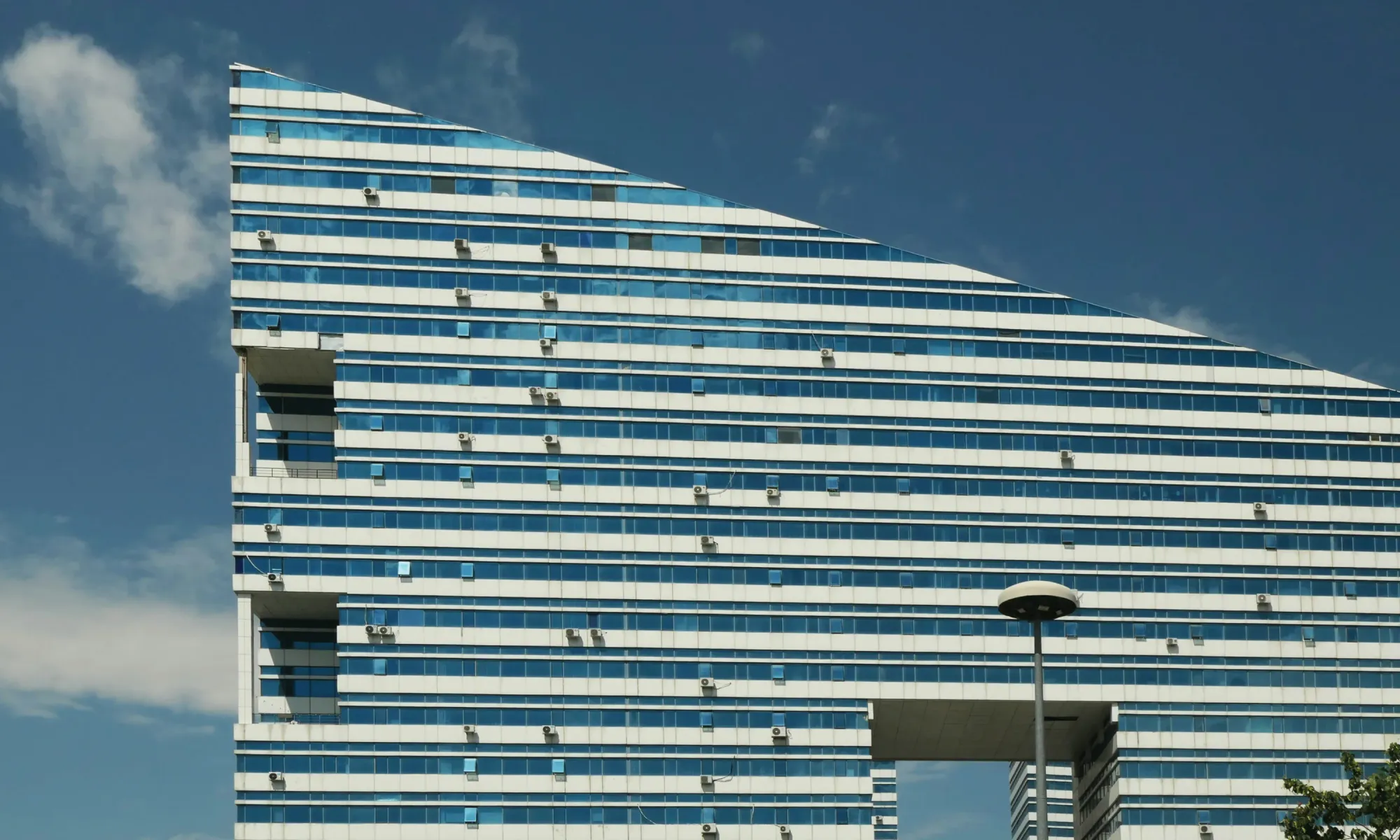 Triangular-topped glass office building with horizontal blue-white stripes and many exterior AC units under a partly cloudy sky in Astana.