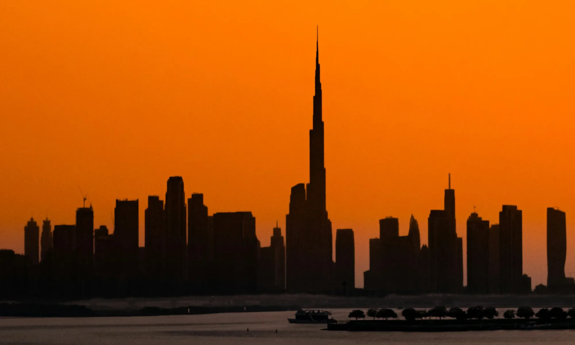 Silhouette of the Dubai skyline at sunset, with the Burj Khalifa towering above all buildings against an orange sky.