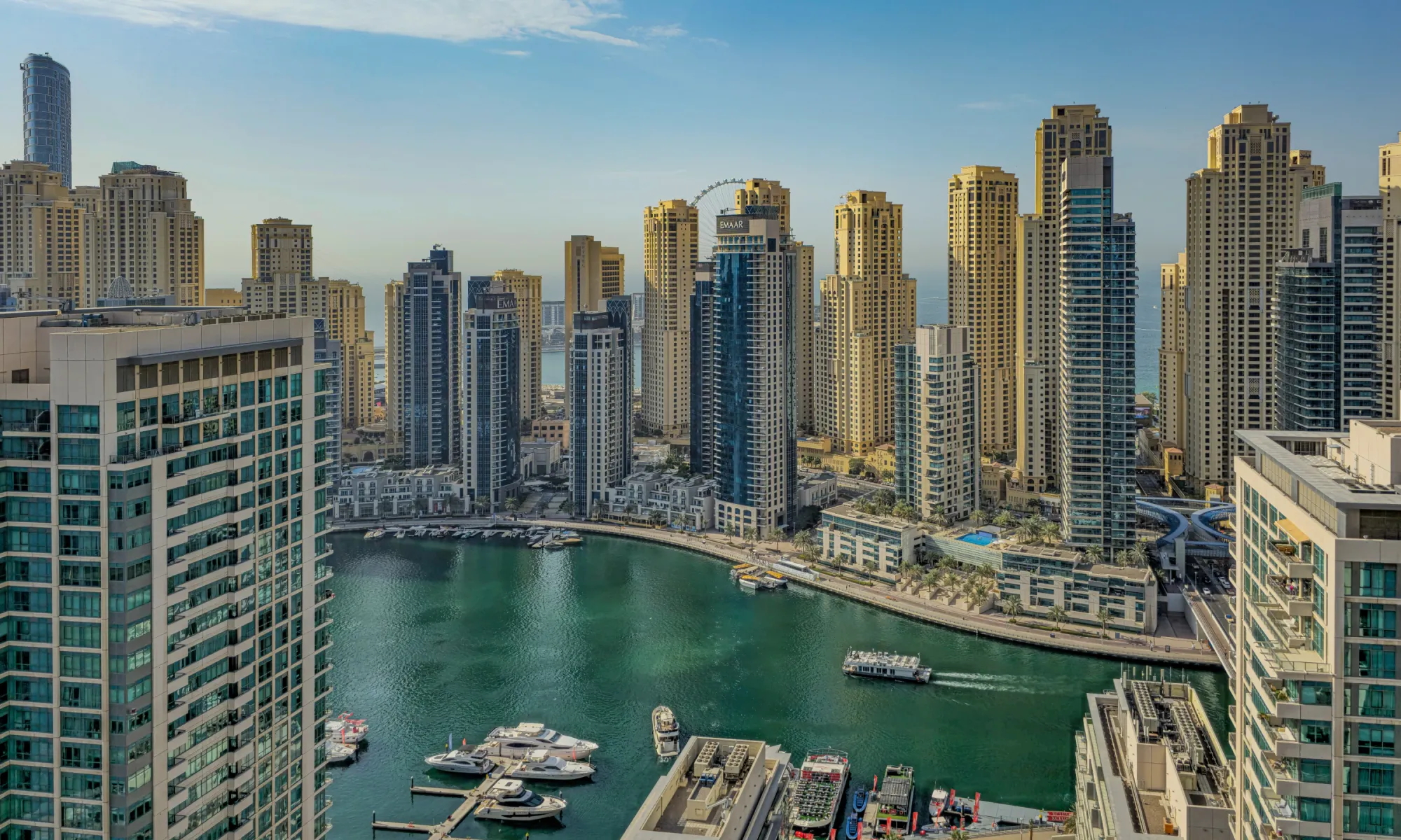 Dubai Marina skyline with high-rise towers surrounding the waterway, boats docked along the waterfront, and a clear sky overhead.