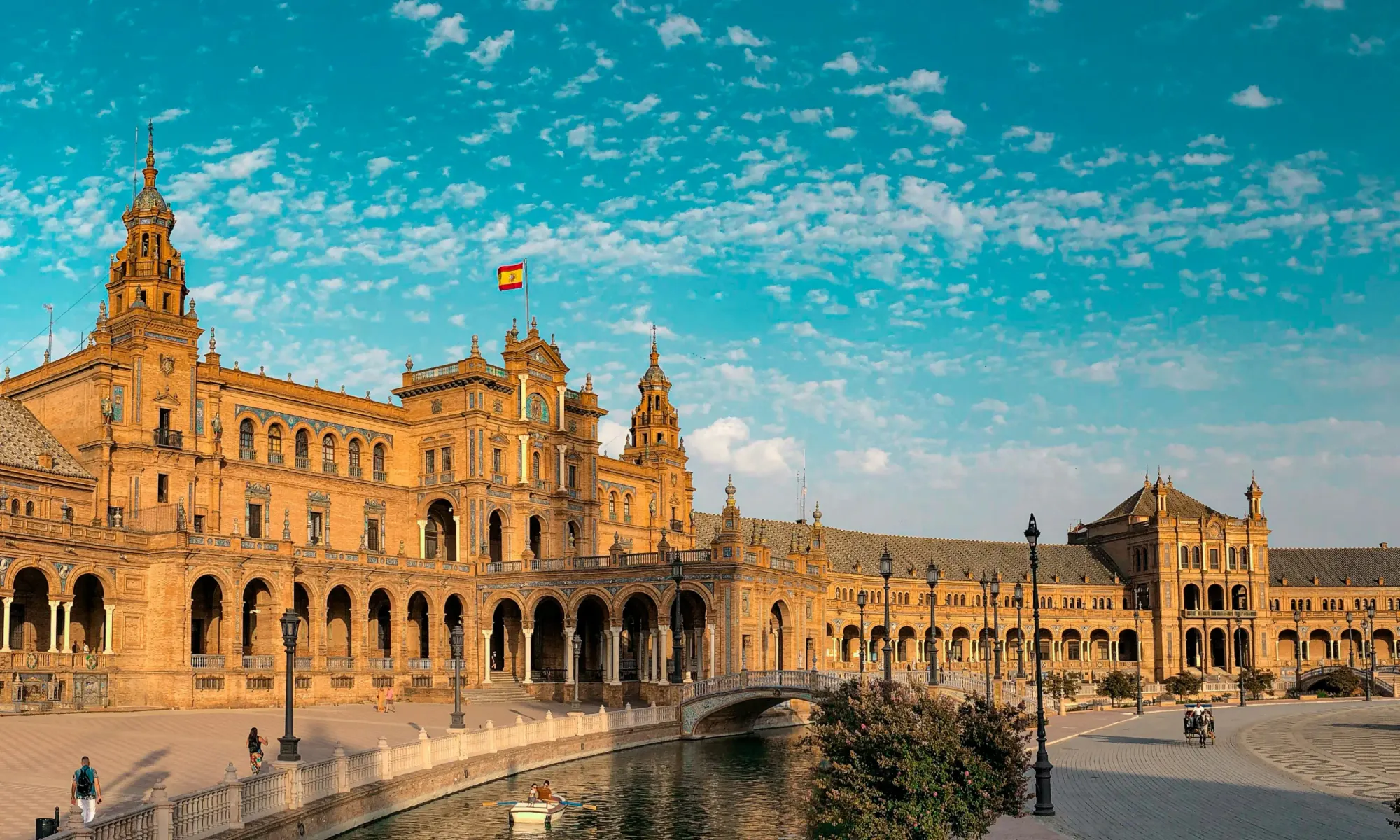 Plaza de España in Seville with arcades and towers, the Spanish flag on the roof, and a canal with a small boat in the foreground on a sunny day.
