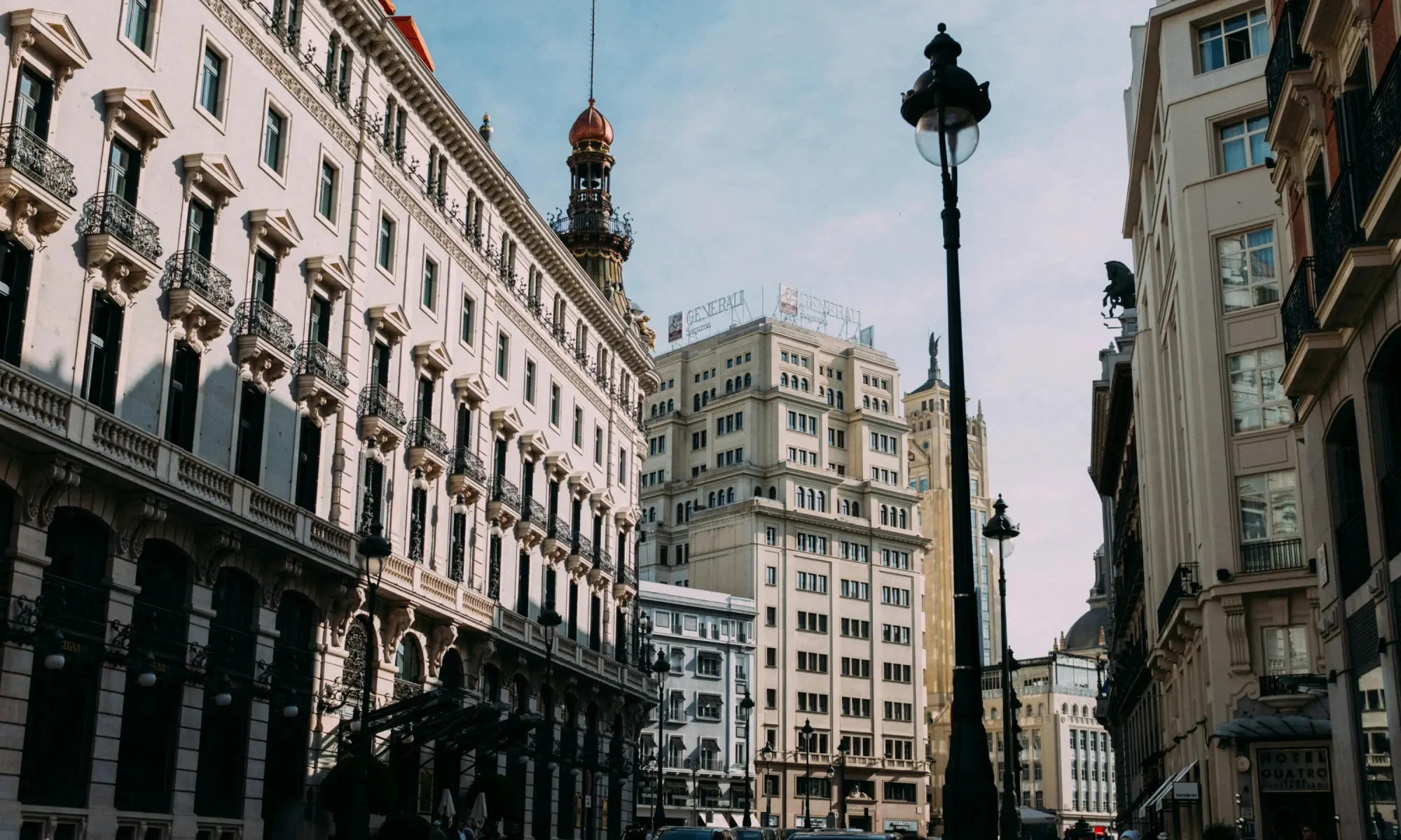 Street view in central Madrid with historic classical-style buildings, Generali sign on a tower in the background, and ornate balconies and lampposts in the foreground.