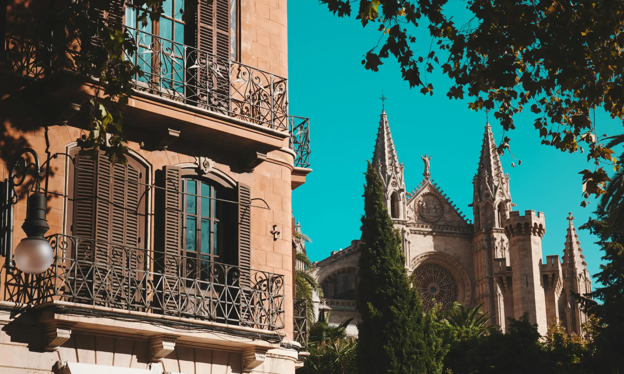 Residential building facade with wrought-iron balconies and shutters, with a Gothic cathedral featuring stained glass and spires in the background in Palma de Mallorca, Spain.