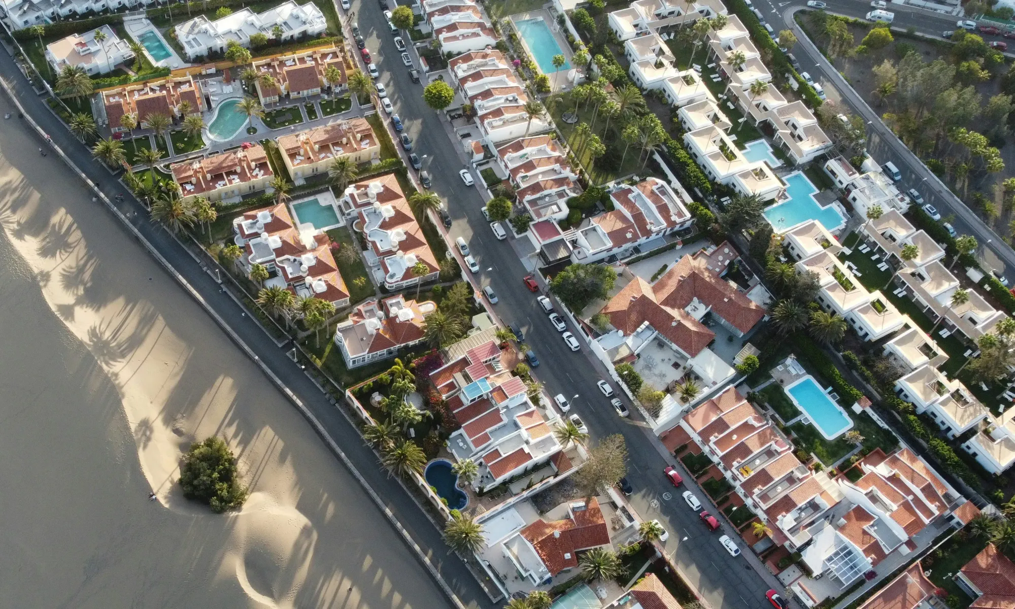 Aerial view of beachfront villas in Marbella, Spain, with red rooftops, swimming pools, palm trees, and a sandy beach on the left.