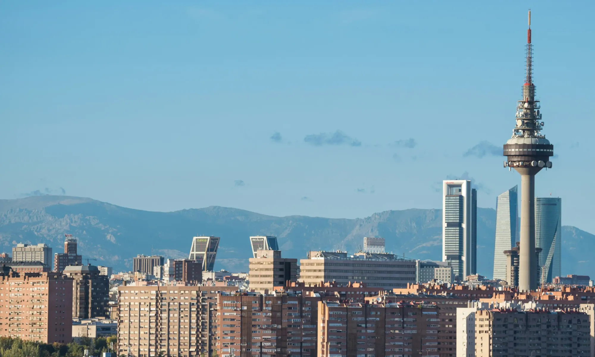 Madrid skyline with Torrespaña communications tower and Cuatro Torres skyscrapers set against mountain ranges in the background.