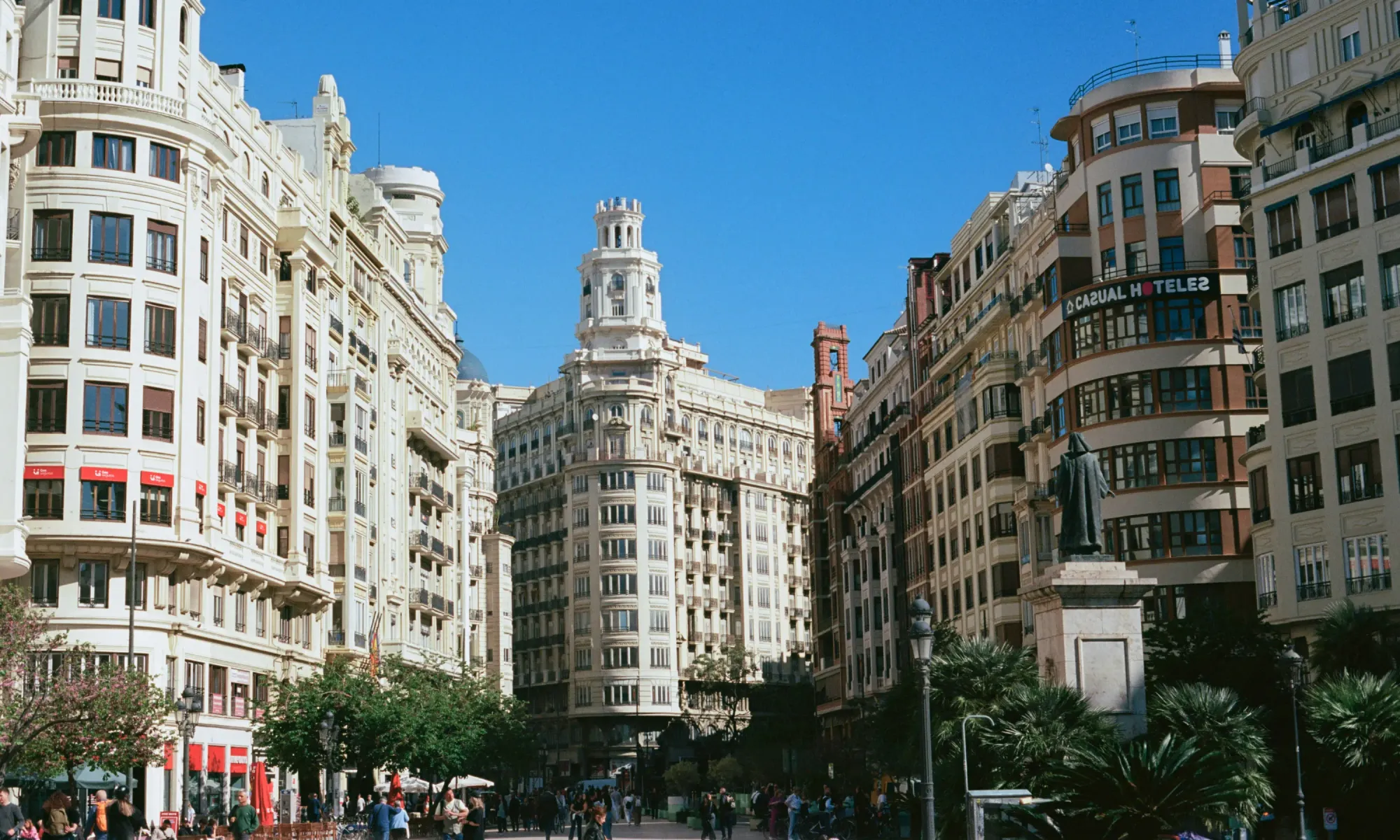 Historic buildings in Valencia’s city center square with ornate facades, a statue, and people walking below on a sunny day.