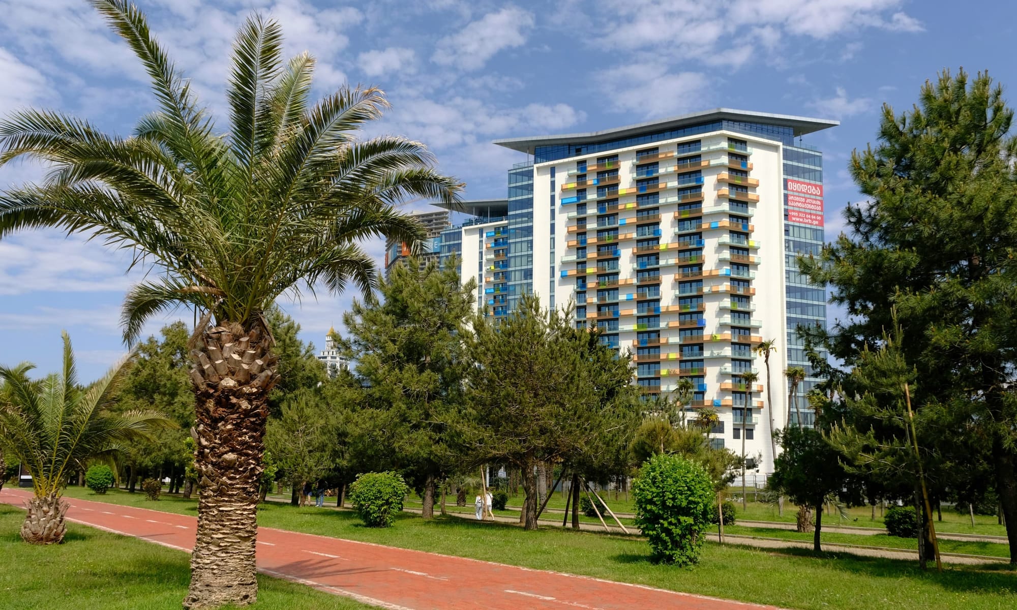 Modern residential tower with palm trees – Contemporary apartment building in Batumi, Georgia, with a colorful facade, viewed from a park with palm trees and walking paths.
