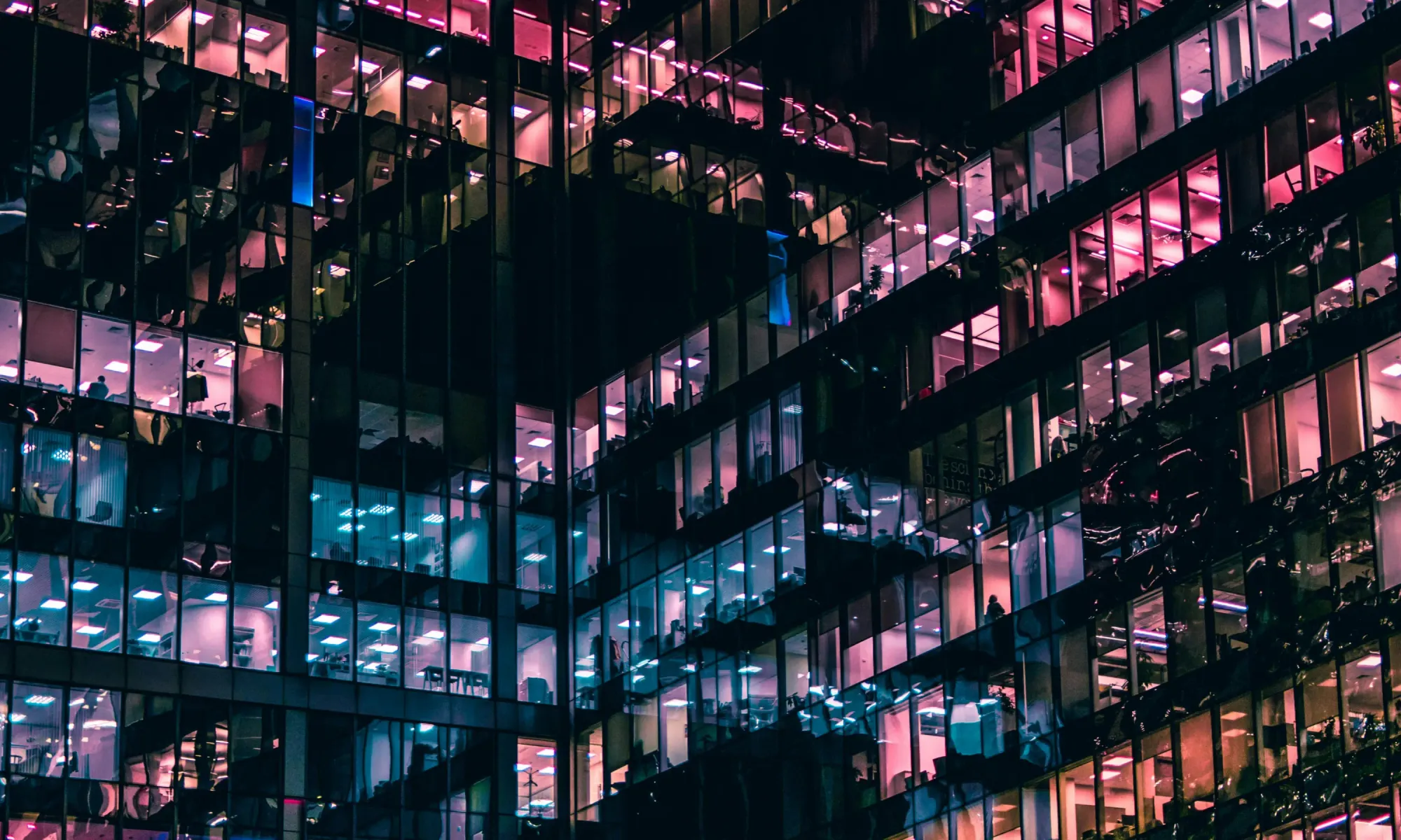 Night view of illuminated office tower windows glowing in pink, purple, and blue tones.