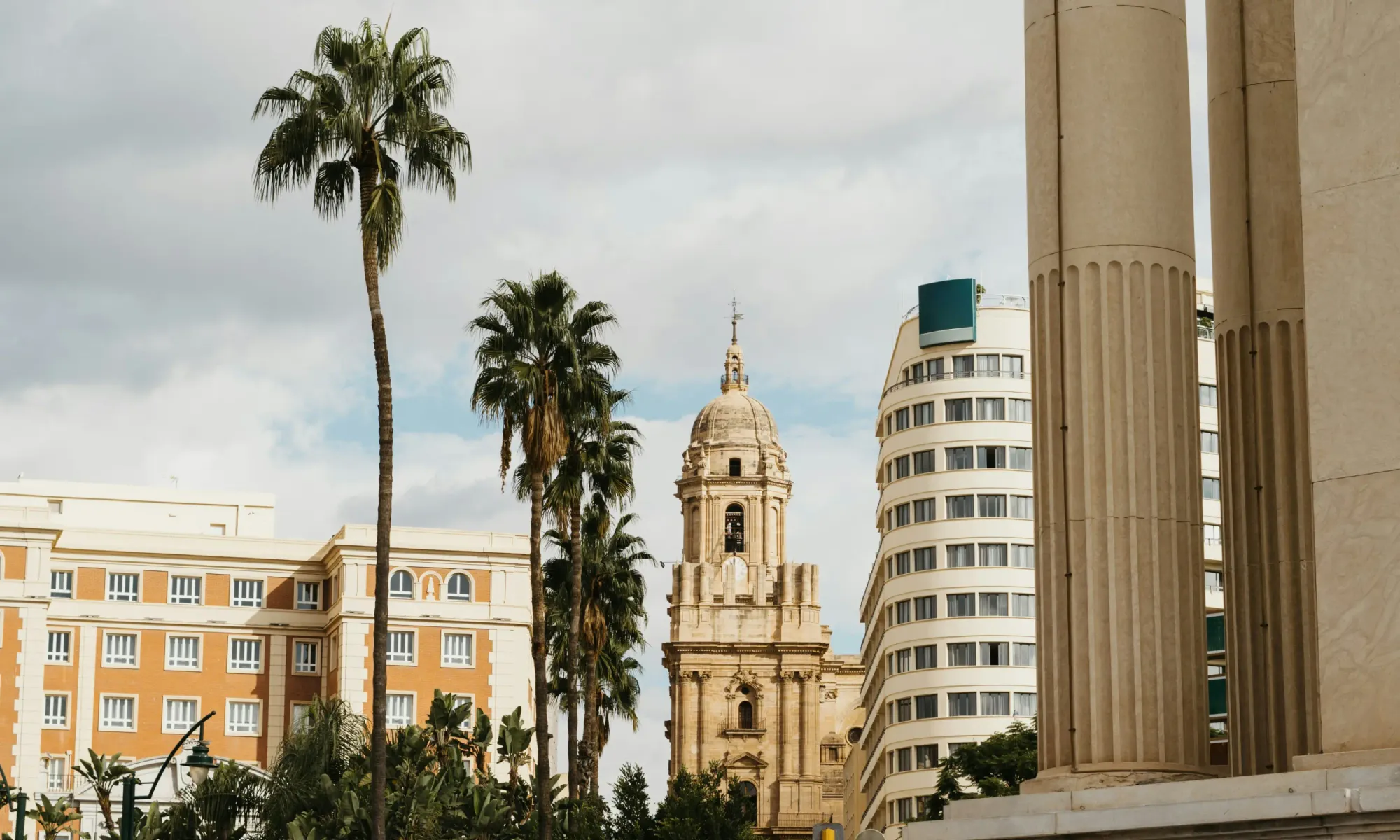 View of Málaga Cathedral with its tall bell tower, surrounded by palm trees and modern buildings in the city center.