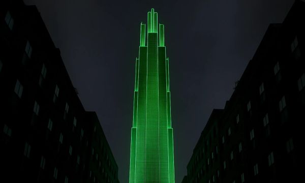 Dark night scene: a tall, tiered skyscraper outlined in vivid green light rises between shadowed mid-rise buildings under a cloudy sky.
