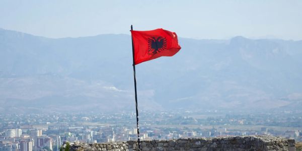 The Albanian national flag with the double-headed eagle waving in front of mountains and a cityscape backdrop.
