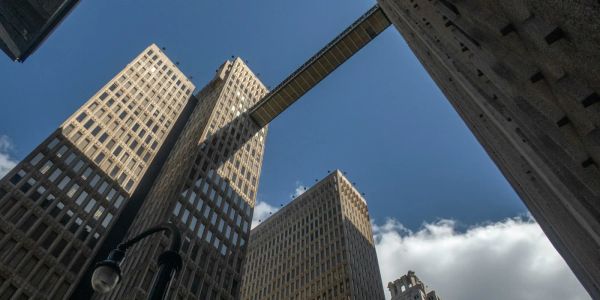 Two tall office towers connected by a skybridge, viewed from below with blue sky and clouds above.