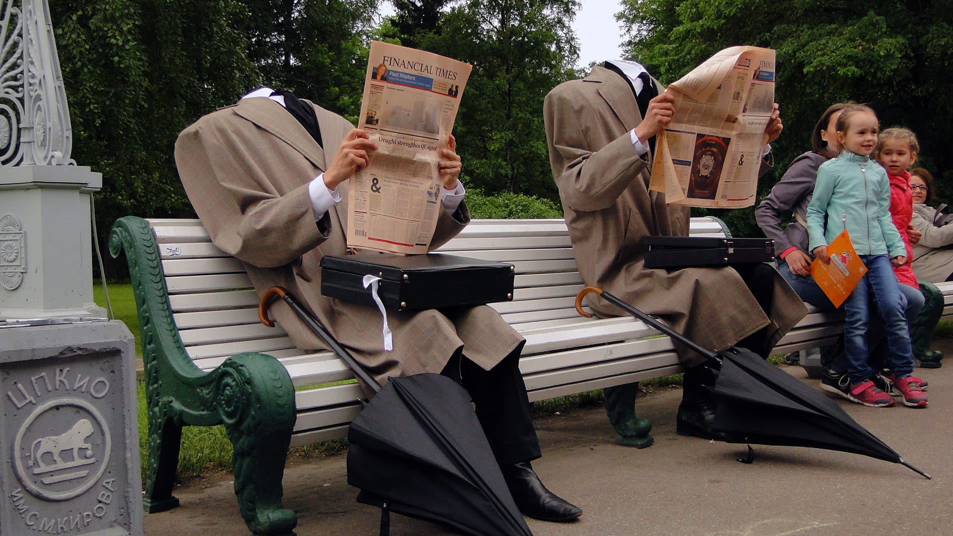 Two people with their heads hidden sit with newspapers on a park bench