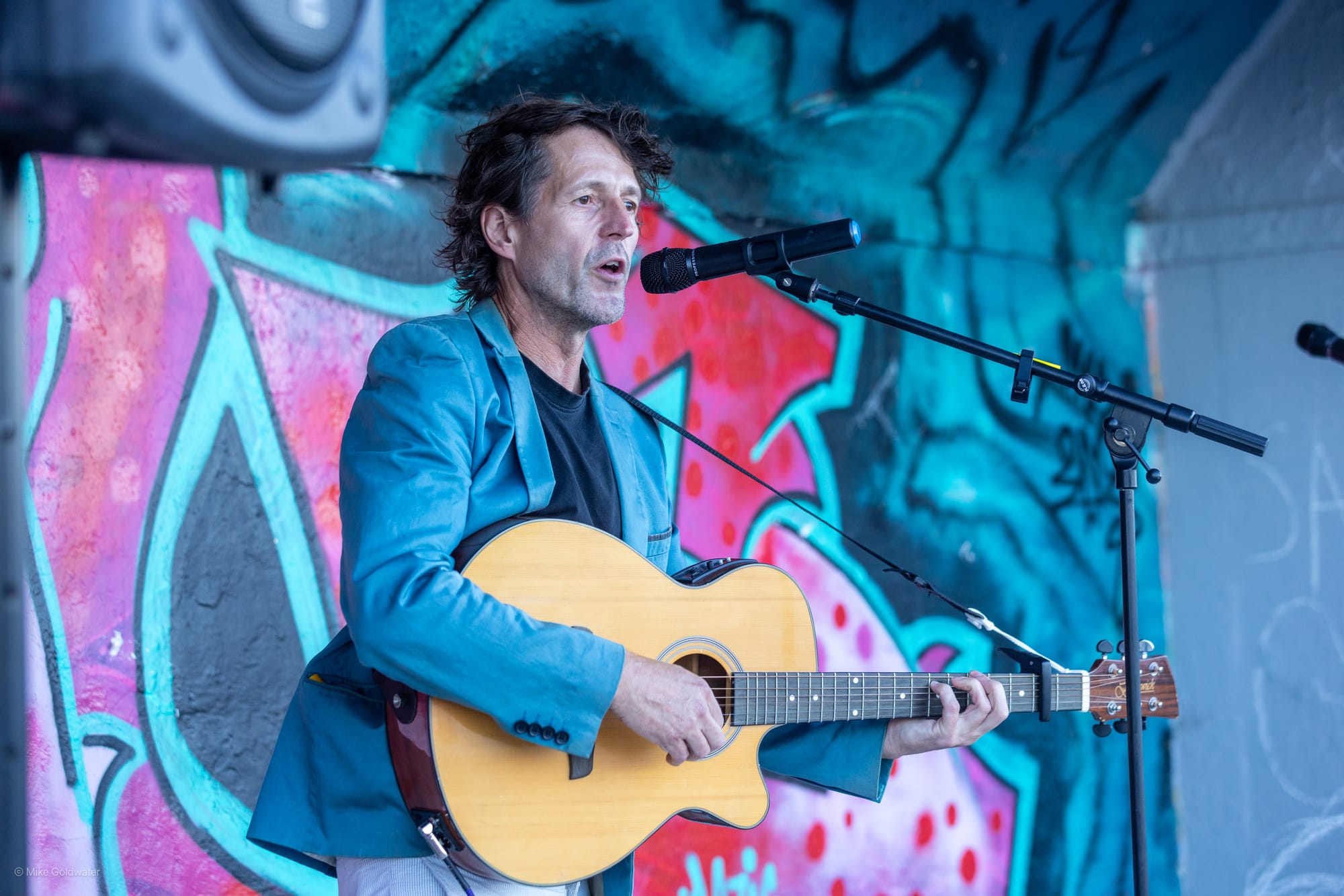 A man with an acoustic guitar plays in front of a wall covered in graffiti
