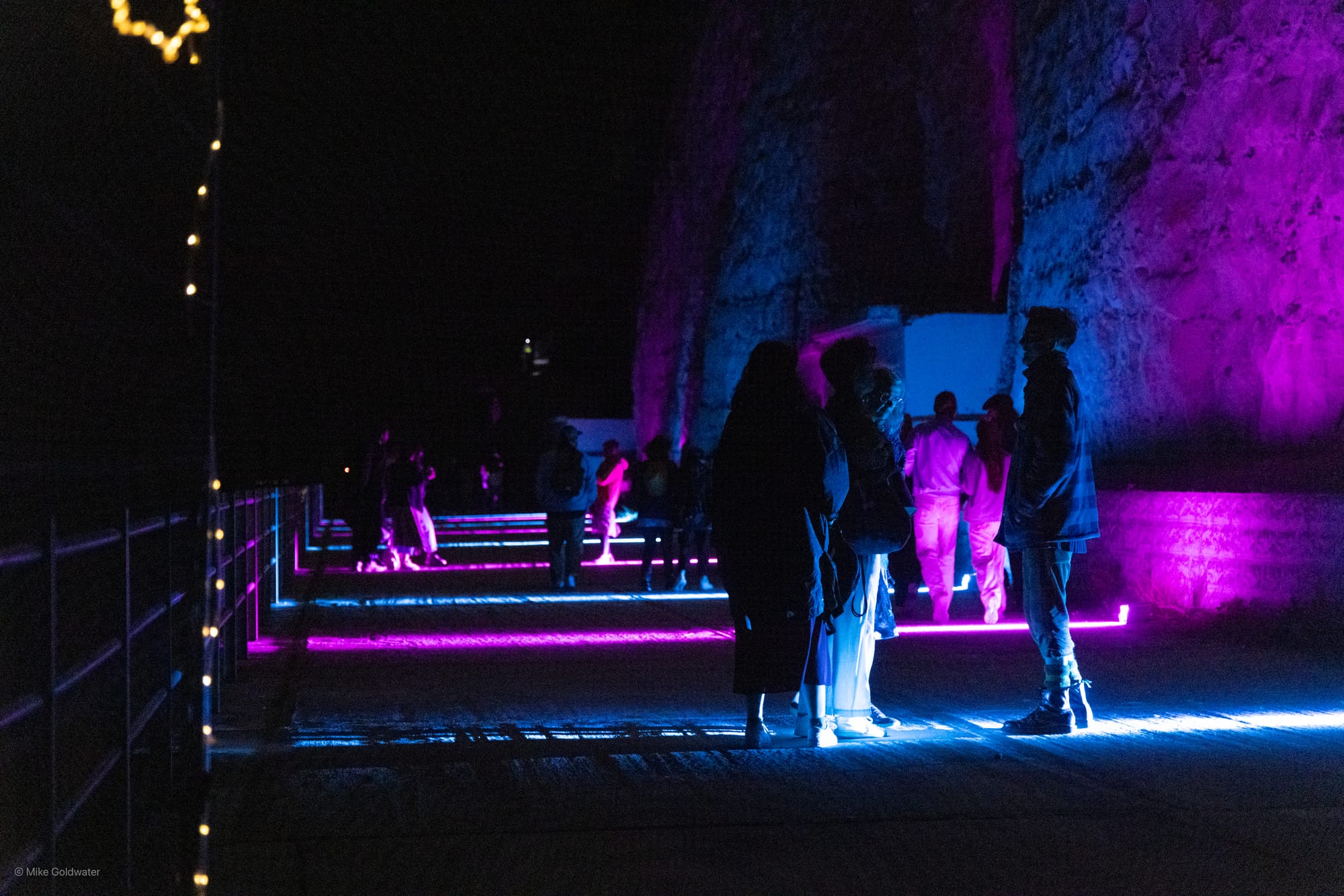 People stand on a promenade next to cliffs lit up purple and blue