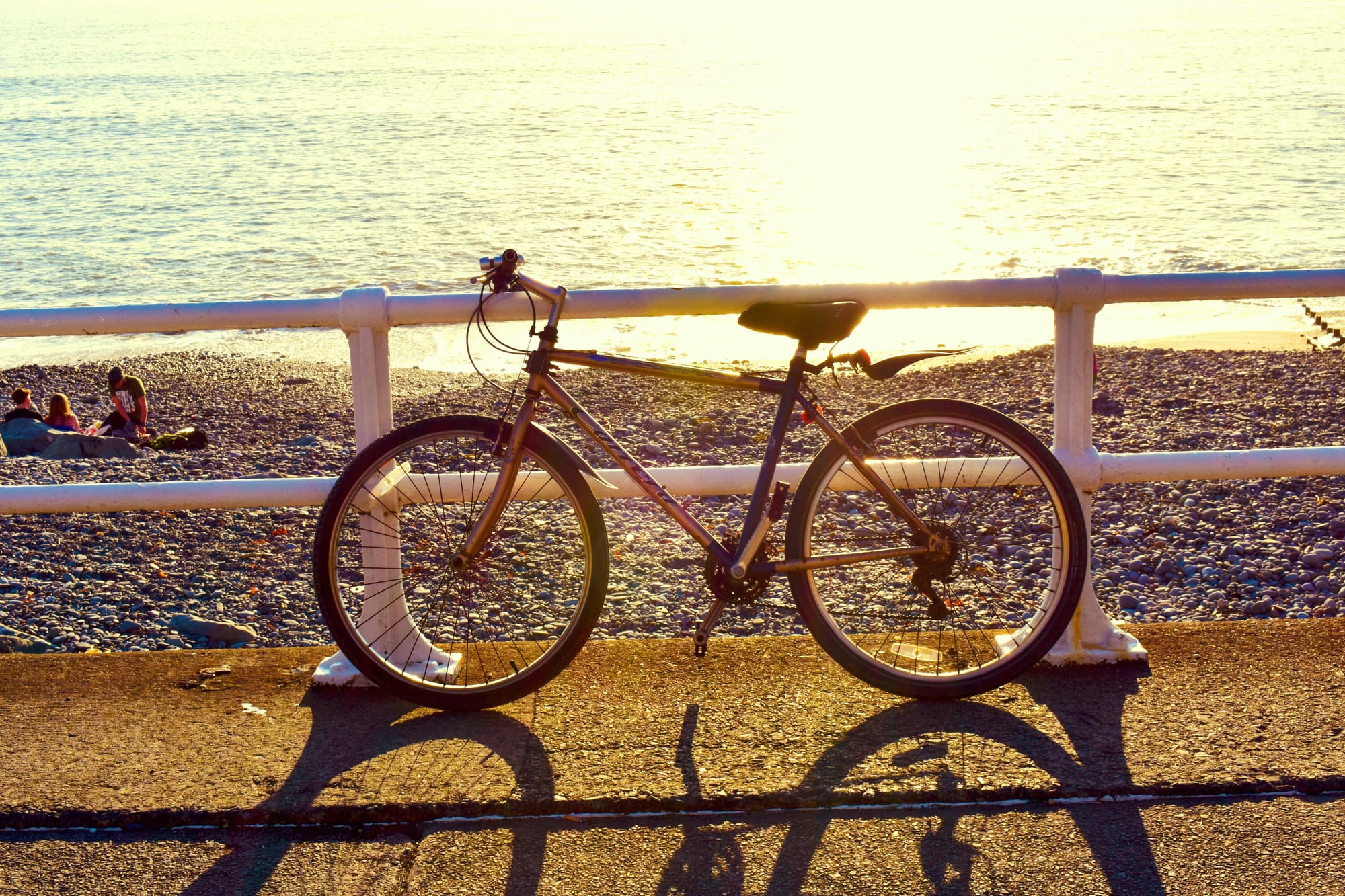 A road bike leaning against rails on a coastal path with the sun and a shingle beach in the background