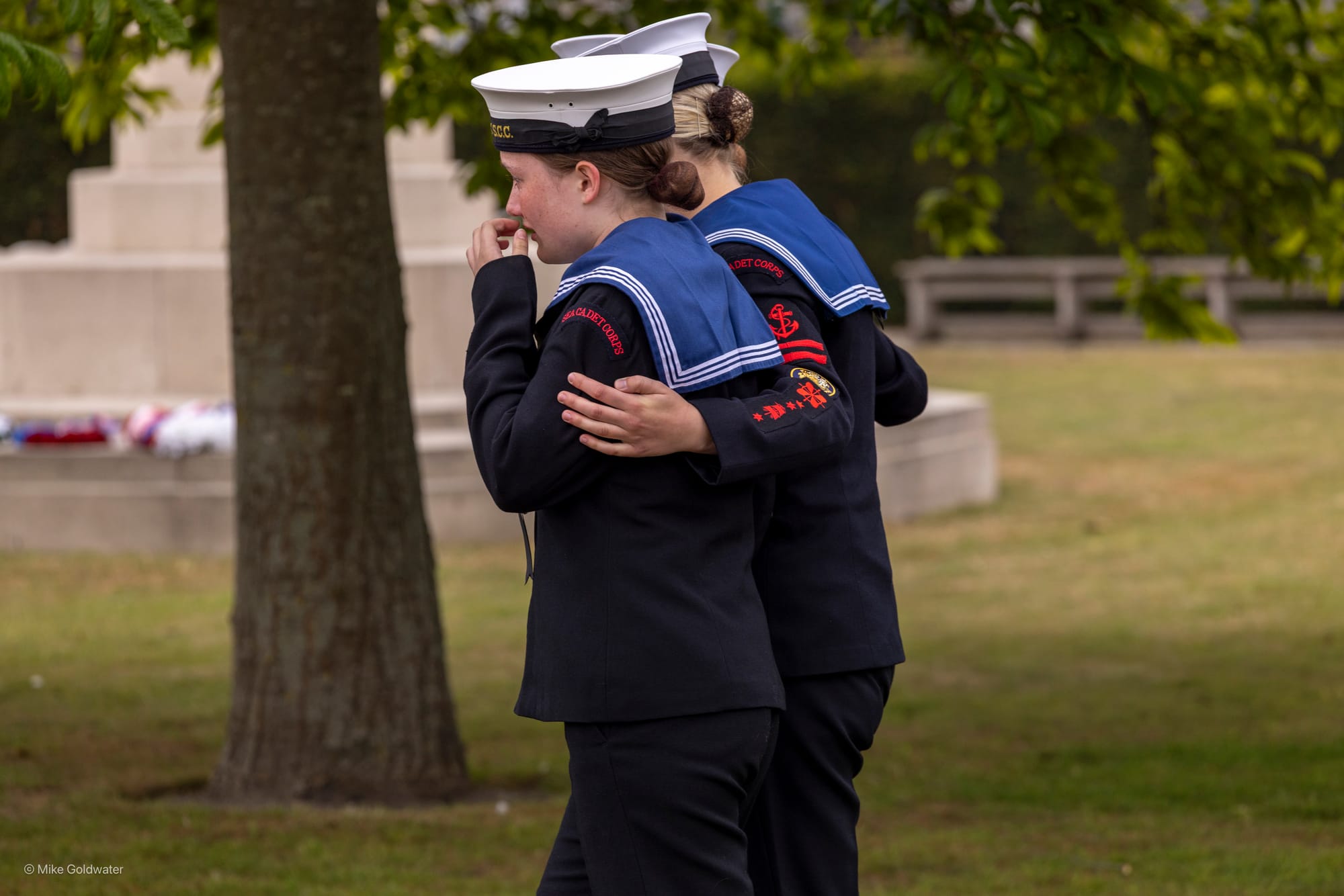 Two teenage girls dressed in sailors uniform, one comforting the other as they walk through a memorial park