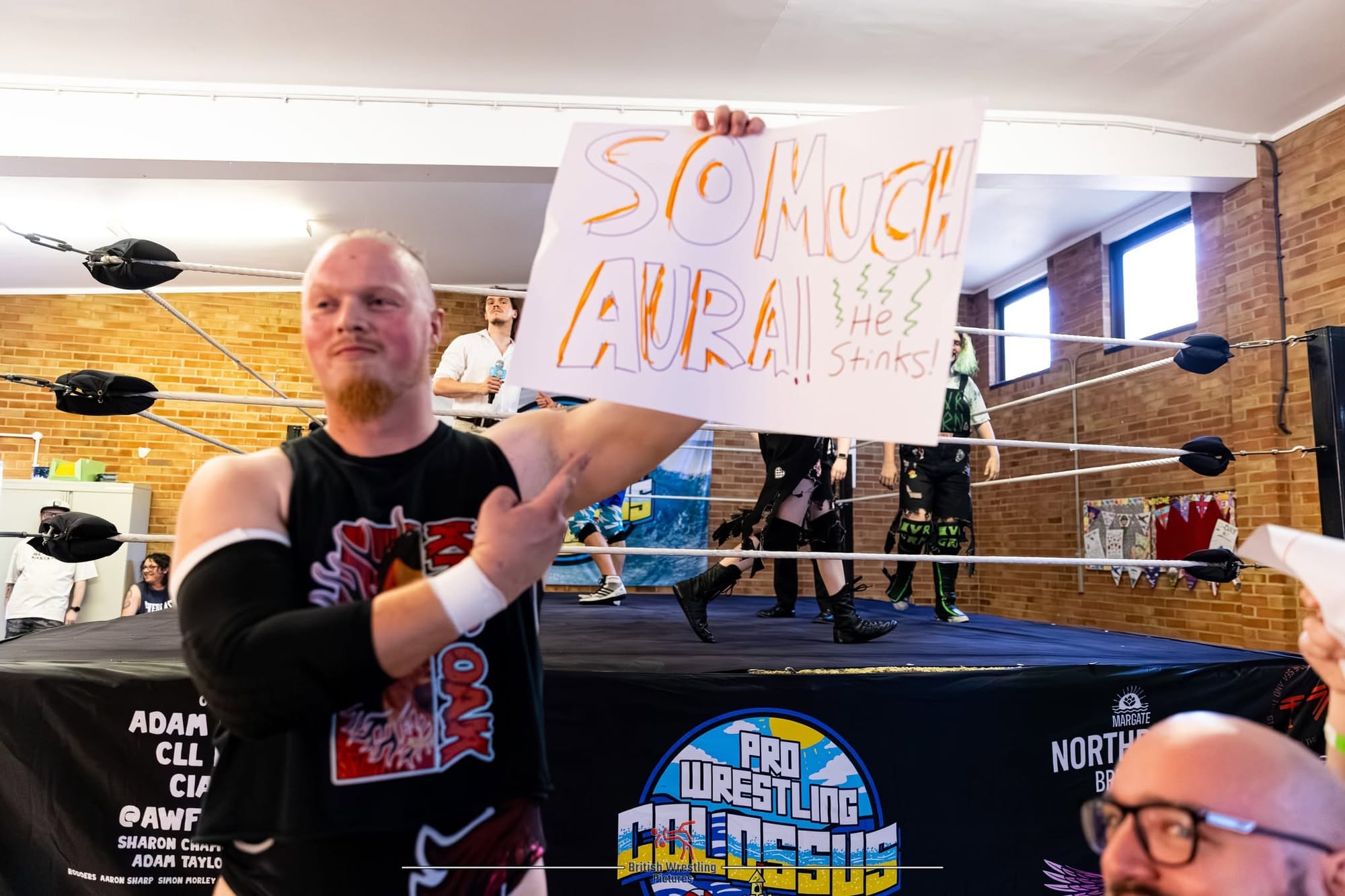 Wrestler holds sign saying So much Aura!! He stinks, in front of the wrestling ring.