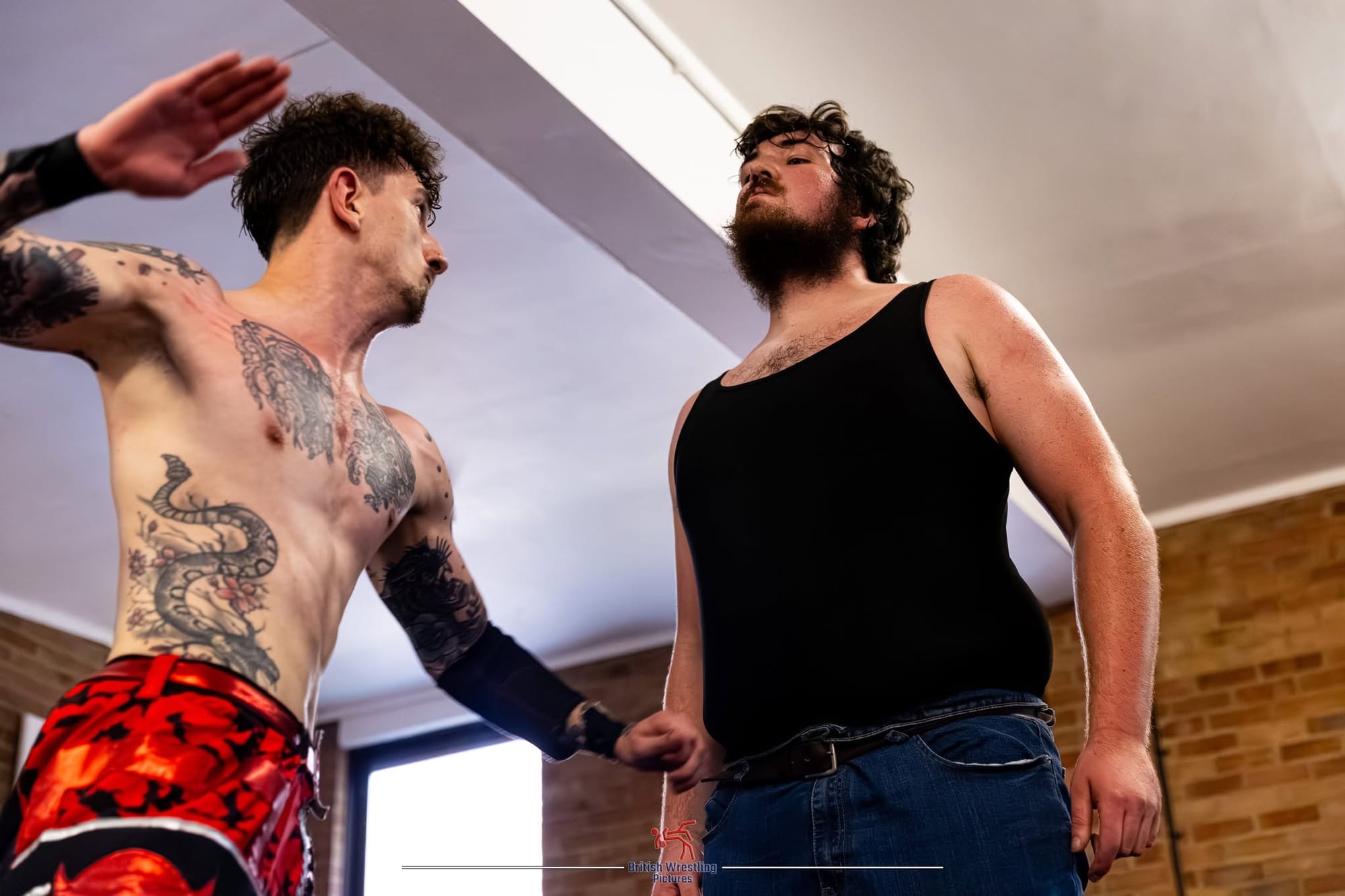 A topless wrestler with tattoos prepares to hit another wrestler who wears a black singlet and braces for the impact. 